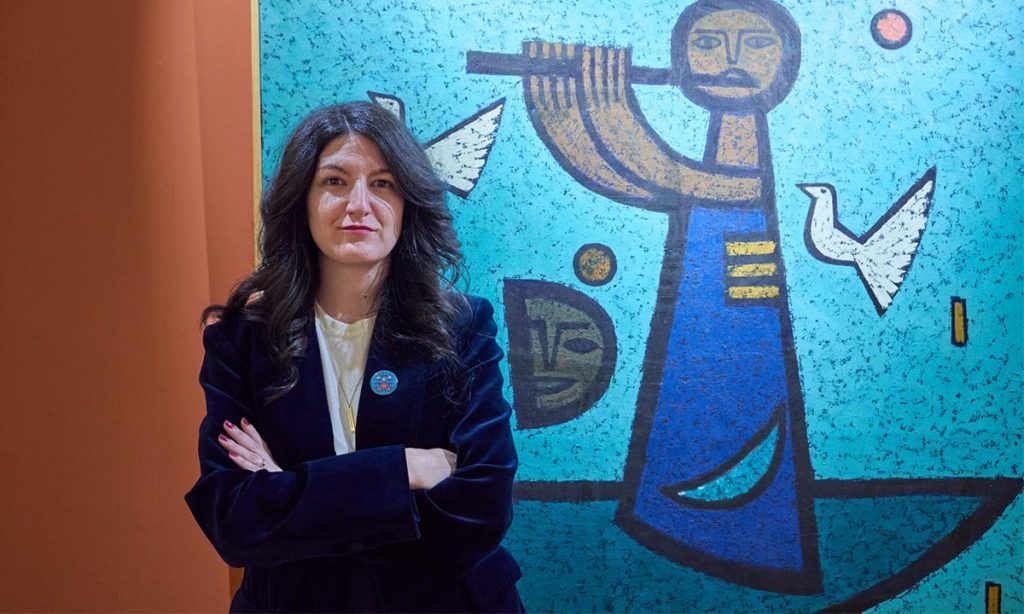 Portrait of a woman with crossed arms in a dark blazer, standing in front of a blue mural in a gallery space.