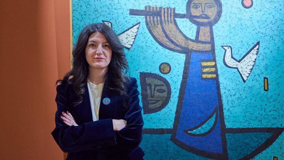 Portret žene sa prekrštenim rukama u tamnom sakou, ispred plavog murala u galerijskom prostoru.
