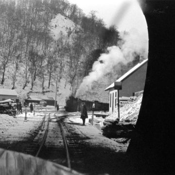 Crno-bela arhivska fotografija: pogled iz tunela na snegom prekrivene železničke šine, mali stanični objekti i parna lokomotiva sa dimom u zimskom pejzažu.
