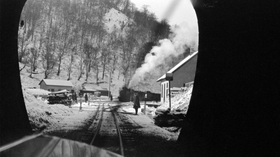 Crno-bela arhivska fotografija: pogled iz tunela na snegom prekrivene železničke šine, mali stanični objekti i parna lokomotiva sa dimom u zimskom pejzažu.