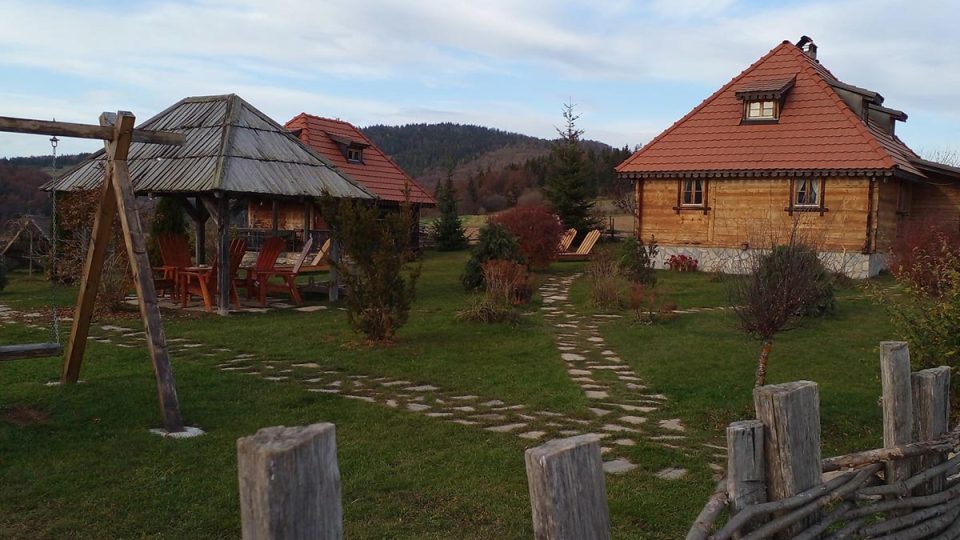 Drvene brvnare i letnjikovac u dvorištu etno domaćinstva na Rudnu, sa kamenim stazama, ljuljaškom i planinom u pozadini.