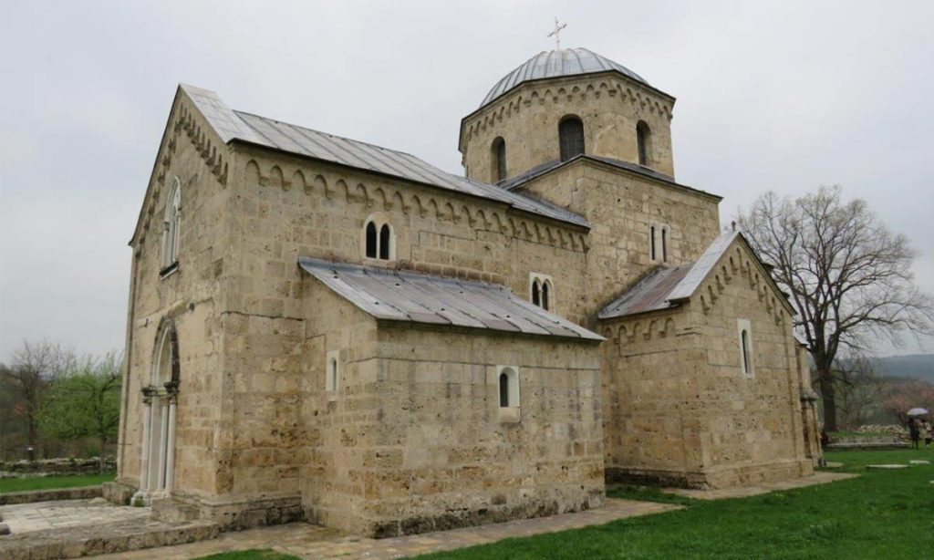 Kamena crkva manastira Gradac sa kupolom i metalnim krovom, na travnatoj porti pod oblačnim nebom.