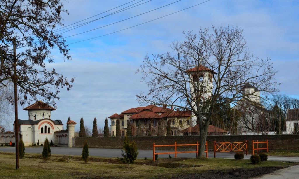 Panoramski pogled na manastir Kovilj: manastirski zid, ulazna kapija i kompleks sa crvenim krovovima iza drveća.