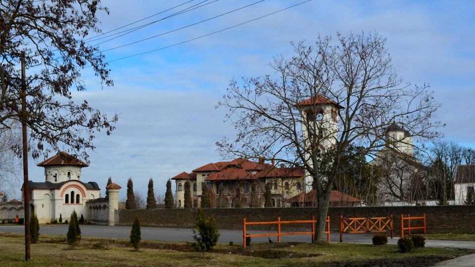 Panoramski pogled na manastir Kovilj: manastirski zid, ulazna kapija i kompleks sa crvenim krovovima iza drveća.