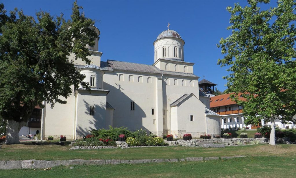 Bočni pogled na crkvu manastira Mileševa u porti, sa zelenim travnjakom, cvećem i drvećem pod vedrim nebom.