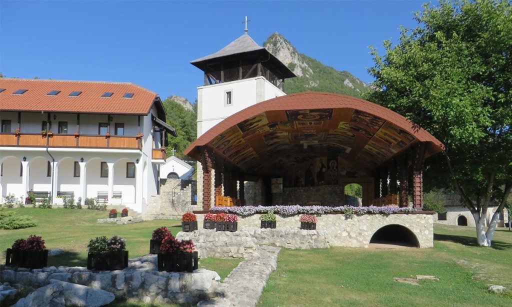 Letnja pozornica u porti manastira Mileševa sa oslikanim svodom, konacima i zvonikom u pozadini.