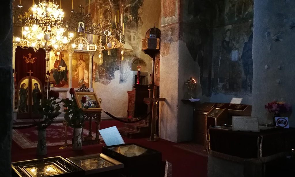 Unutrašnjost crkve manastira Mileševa sa ikonostasom, kandilima i zidnim freskama u polumraku.