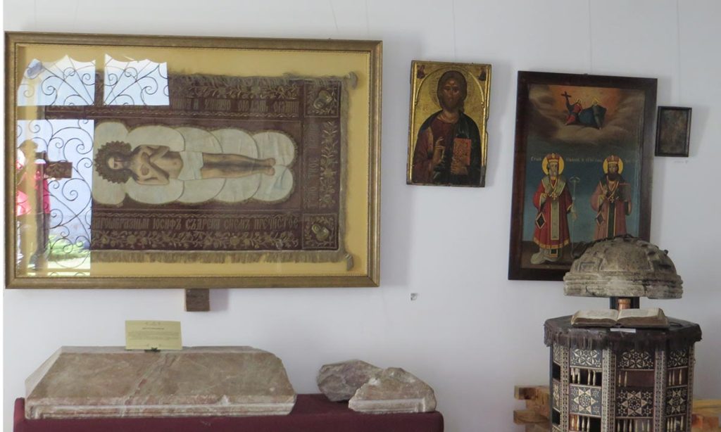 Postavka manastirske riznice u Mileševi sa ikonama na zidu, izloženim liturgijskim predmetima i knjigom na drvenom postolju.