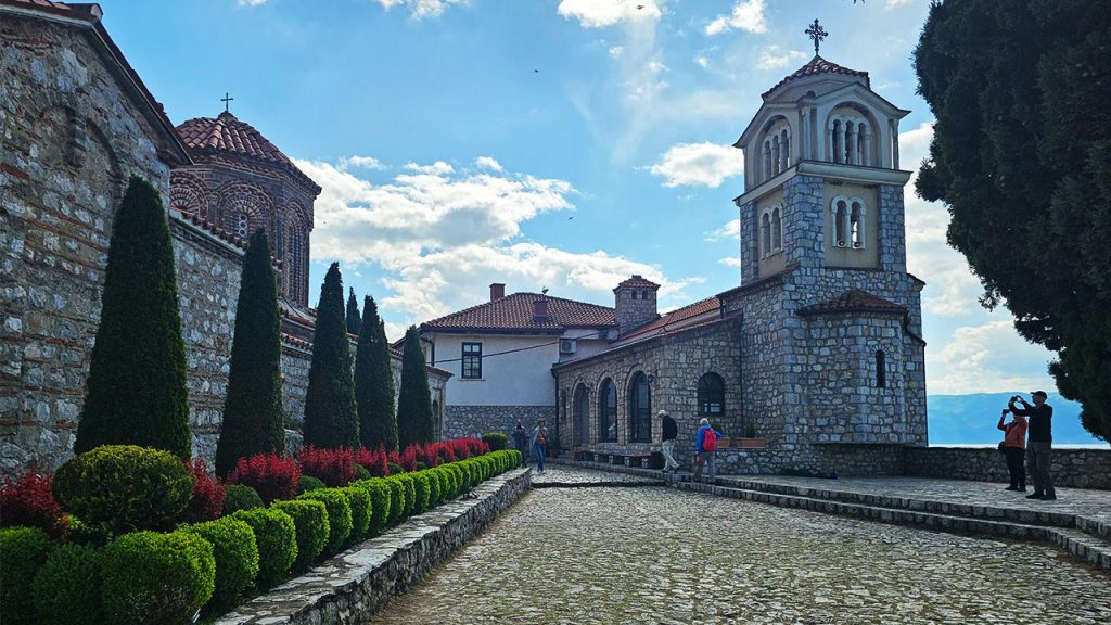 Dvorište manastira Svetog Nauma na obali Ohridskog jezera, kameni zvonik i crkva uz čemprese i posetioce.