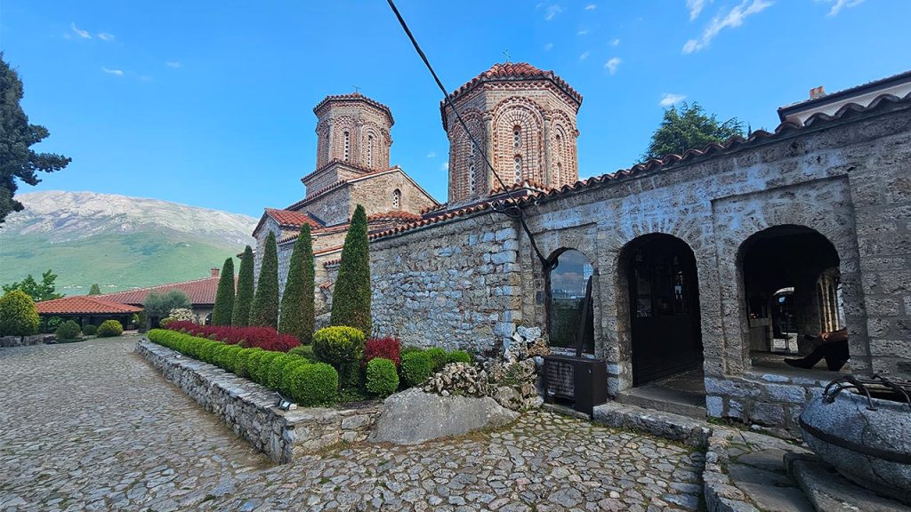 Pogled na manastirski kompleks Svetog Nauma: kameni trem sa lukovima, uređene živice i crkveni tornjevi pod vedrim nebom.